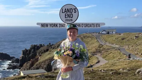 A picture of Ella Fryer holding a bouquet of flowers in front of a Land's End sign