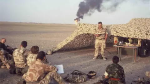 Combat Stress Mark Christian standing in front of a group of soldiers who are sitting on a dusty barren plain while on deployment in Iraq. A camouflage tent has been set up behind them, under which is a table with a Christian cross on top. In the distance there is a fire with black smoke billowing into the sky.