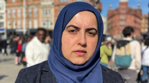 A head and shoulders shot of Neghat Khan, leader of the city council, wearing a blue hijab stood outside with crowds behind her 