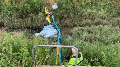 Environment Agency officers at Dovers Brook trying to clear the oil spill -  men are in the water with a long blue tube and boards