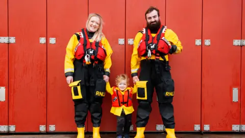 RNLI Emily Craven, who has blonde hair, and Tom, who has brown hair and a thick broan beard, smile at the camera holding the hand of a toddler with curly blonde hair. All three are wearing yellow and black full body waterproofs and red life-vests. 