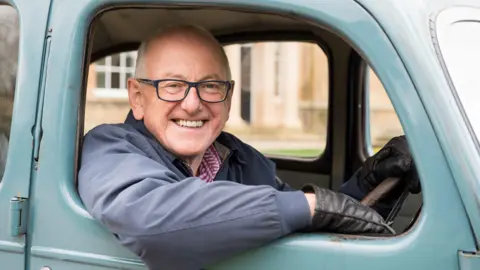 A man with short grey hair and glasses smiles from the driver's seat of a blue vintage car.