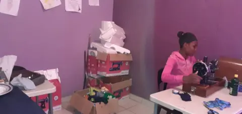 LiDAR Community Foundation's young pink woman sitting at the table with sewing machine in the room with purple walls. She was surrounded by cardboard boxes, sewing supplies and some posters attached to the wall.