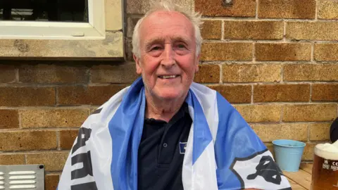 Kate Bradbrook/BBC A man smiles at the camera as he sits in a beer garden. Behind him is a yellow brick wall. He has grey hair, a blue and white flag wrapped around his shoulders and he is wearing a navy polo shirt underneath.