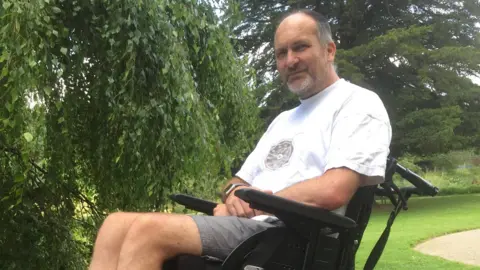 Julie Heath A man in a manual wheelchair sitting on a lawn next to a path with mature trees behind him on a bright day