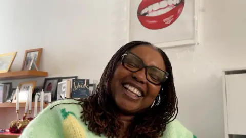 BBC Yvonne Johnson laughing sat in her living room in her wheelchair, with a portrait of her lips behind her.