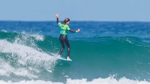 Boardmasters A person wearing a green top and black wetsuit balances on a surfboard while riding along the crest of a small ocean wave. White foam splashes behind the surfboard, with deep blue water and a clear sky stretching across the background.