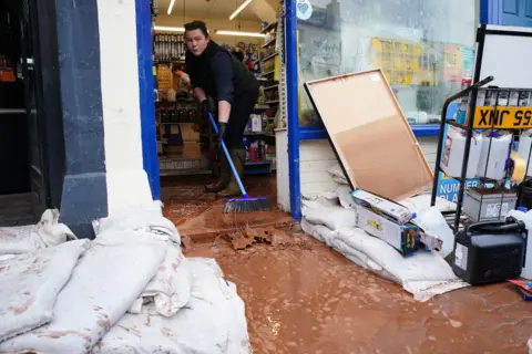PA Media Woman sweeps brown muddy water out of a shop door with a gap made in sandbags over the front step