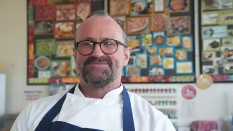 Daniel Smith inside a classroom wearing chef whites and a blue apron. He has a pair of black framed circular glasses on and is looking off camera mid-speech.