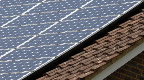 Generic image of solar panels on the roof of a building. The roof also has brown tiles on it.