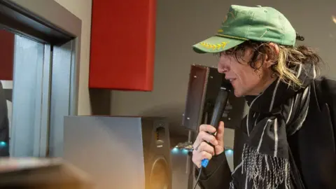 Hereford Cathedral School Justin Hawkins has long curly brown hair and is wearing a green cap and black coat with a black and white scarf. He is in a recording studio and is standing in front of a large glass window, holding a black microphone and speaking into it.