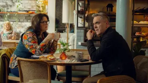 Netflix Ruth Jones wearing a floral top with a dark green waistcoat. She sits at a cafe table holding a coffee cup, as she looks to a tense James Nesbitt who sits opposite her in a black jacket and beige trousers.