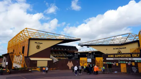 The exterior of Molineux Stadium. It is a Light yellow, dark yellow and black stadium. There are visitors stood outside the entrance which shows the Billy Wright stand and Sir Jack Hayward stand.