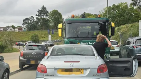 A lorry with a car parked in front of it. A woman is stood by the car with her back to the camera. A number of cars are parked by the side of the road around them.