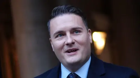 Wes Streeting leaves a building and he appears to be talking. He has short dark hair and wears a navy suit with a blue shirt and blue polka dot tie. 
