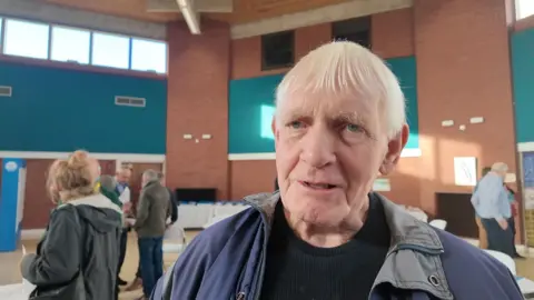 A man with white hair wearing a blue jacket stands in the middle of the exhibition about the proposed reservoir