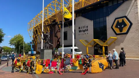 Floral tributes, flags and shirts are placed in a circle at the foot of the Billy Wright statue at the Wolves ground. A yellow fence has been erected around the statue, with shirts and scarves of different colours draped over it. In the background is the outside of the football stadium, with the words 'Billy Wright stand' on the wall. There is also the yellow and black wolf badge of the Wolves logo on the side of the stadium.