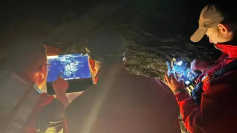 Three people at night crouched toward the ground. Their backs are to the camera. They are looking at screens that show drone thermal images.