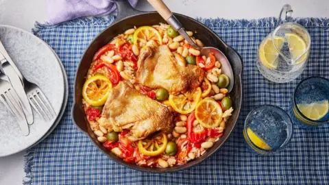 A round pan of Mediterranean chicken with tomatoes, beans, rice, olives and lemon slices is in the middle of the image. It is sitting on a table on top of a blue and white check tablecloth. There is a pile of light blue plates on the left hand side with silver cutlery placed on top, and a jug and two glasses of water with lemon slices in it, to the right.