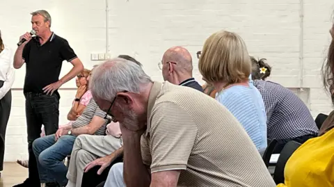 A photograph of a row of seated people looking up at a man at the far end of the row. The man is holding a microphone with one hand and the other hand is on his hip. He is speaking into the microphone. A older man can be seen in the foreground resting his head in his hands.