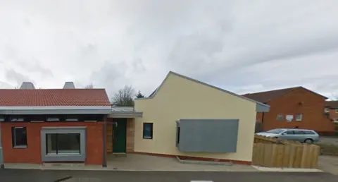 Google A single-story brick-built house, with a brick coloured section on the left and painted cream-coloured section on the right. There are two large picture windows - one of which has a metal shield over it.