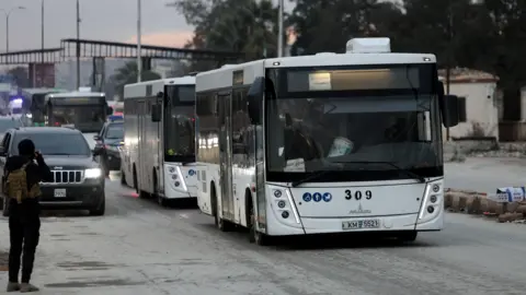 Reuters A person in black is seen taking photos of two white buses, carrying members of the Syrian Democratic Forces (SDF) out of Aleppo