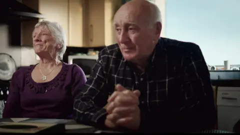 Firecrest Films A tearful elderly woman with short grey hair and wearing a purple knitted looks off to her right, Her husband, who is bald with grey hair at the sides, sits with his hands folded. He is wearing a checked shirt and is also looking off to his right. The couple are sitting at a kitchen table.