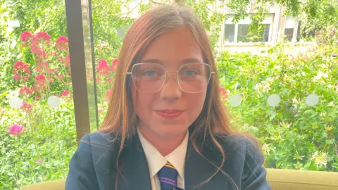 BBC/Carla Fowler Lexi seated indoors in front of a large window. She is positioned centrally in the frame and is facing directly toward the camera. She is wearing a dark-colored blazer over a white shirt and a striped tie. She also has round, thin-framed glasses and long hair that falls over the shoulders.
