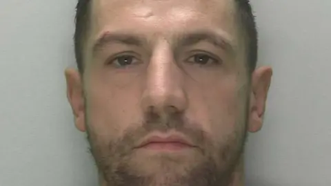 Gloucestershire Constabulary Scott McGuinness looking at the camera in a custody mugshot. He has short dark hair and scruffy stubble