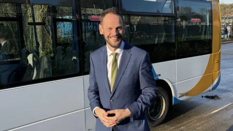 BBC Simon Lightwood wearing a navy blue suit and gold tie and white shirt standing smiling with his hands clasped together in front of a white bus.