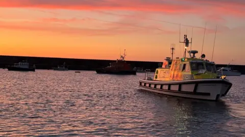 St John Ambulance & Rescue Service, Guernsey The ambulance boat on the water in Alderney at sunset, with other boats in the background, amidst pink and orange skies.