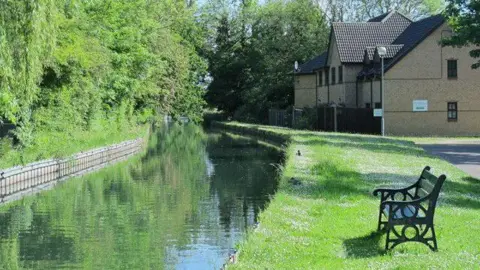 Mike Quinn/Geograph River curving round to the left with hedge on the left bank behind log roll, and grass on the right with a dark-coloured bench looking towards the river. There are new houses in the background.