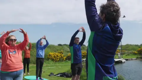 Neil standing on grass on yoga mats next to a lake with a group of other swimmers with hands above their heads