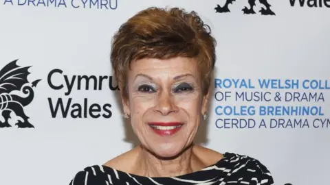 Iris Williams smiling at the camera. She wears a black and white top. She is standing in front of a white background with Cymru Wales, in black writing, and Royal Welsh College of Music & Drama, in blue writing, behind her.