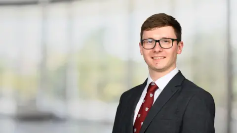 Savills Dan smiling down the barrel of the camera. He's wearing black-framed glasses, a grey suit, white shirt and red tie. He has short brown hair. 