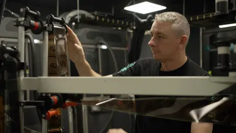 A man with silver hair adjusts a ruler over which film is looped