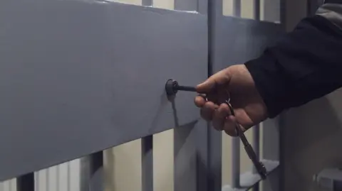 Getty Images A stock image showing a grey metal barred door with a hand putting a key into a lock on it. The person's arm can be seen and they are wearing a dark long-sleeved jacket.