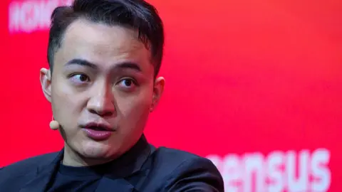 Justin Sun in black blazer and black T-shirt, with small beige microphone and ear piece in front of red background