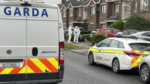 A still image of two forensics investigators at a distance in white suits approaching the property in Malahide. There is a white van with garda written on it in blue writing on the left, and on the right is a white garda car with reflective stripes on the back. In the distance are a number of brown brick semi-detached homes.