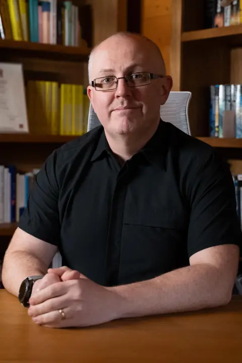 Dan Coughtrey Dean is wearing glasses sits at a wooden desk with hands clasped together, facing the camera. He is wearing a black polo top and watch. He is bald. Behind him are floor-to-ceiling wooden bookshelves filled with books and framed documents, giving the room the appearance of a personal library or office.