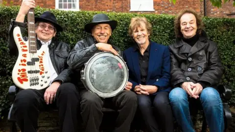 Payley photography Four members of The Zombies band pictured sitting in St Albans in front of a hedge, two holding their instruments - an electric guitar and a drum. They are smiling cheerfully at the camera.