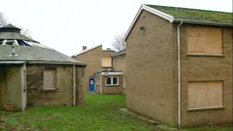 A collection of building boarded up with a blue door in the distance and grass betwen them.