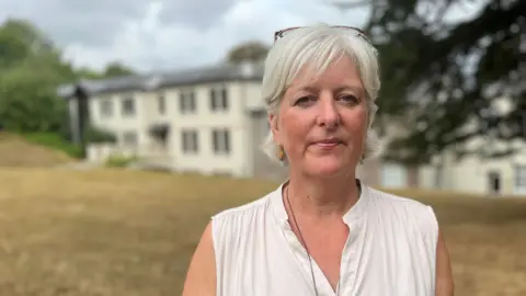Caroline Voaden in a sleeveless cream top against a blurred background of tree foliage and offices at Follaton House in Totnes