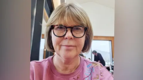 LICAF Julie Tait, founder and festival director at The Lakes International Comic Arts Foundation (LICAF). She has blond hair in a short bob and round black-rimmed glasses. She is wearing a pink jumper and looking at the camera with a slight smile.