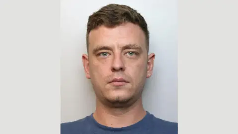 A police mugshot of Kieran Hutchinson. He is wearing a dark blue t-shirt and is standing against a grey background. He has short brown hair which is longer on the top, light blue eyes and a blank expression.