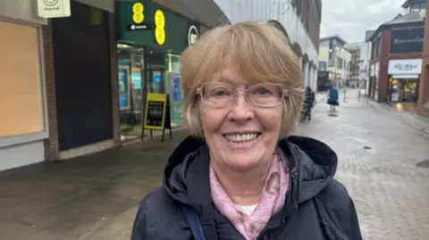A woman with short, sandy blonde hair stands in a high street with a pedestrianised street and shops visible in the background. She is wearing a navy raincoat and glasses and smiles at the camera.