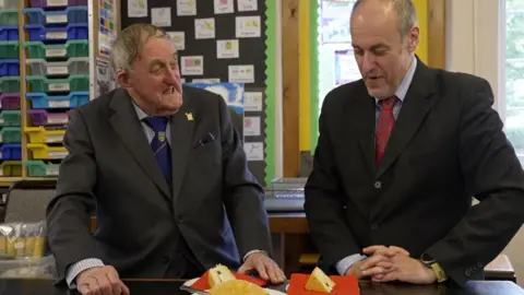 Dr. Neville Brown was given his son - Dr. Sitting in a smart gray suit smiling on deril brown (right) (left) in a pinstrip black suit with a red tie, with a piece of cake in front of them.