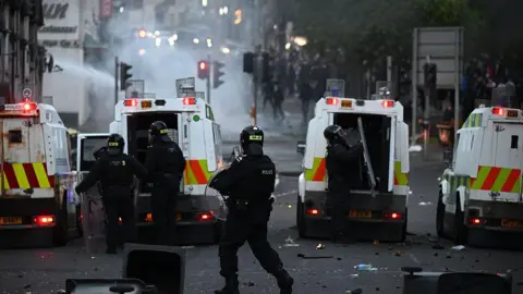 Four white, yellow and red police vans face towards a crowd, away from the camera, as smoke as water appear between the vans and the crowd. There are four people wearing black riot gear holding clear round shields. Smashed glass and other bits of rubbish can be seen on the floor behind the police vans. 