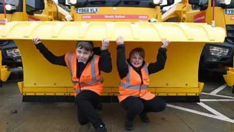 Two school boys wearing hi-vis jackets are crouching under the plough of a large yellow gritter. They have their arms raised and are holding the gritters. A red sign on the gritter announces its name as Ready Salted.