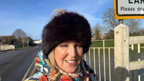 John Devine Angela Lake ahs blonde hair but is wearing a large black fur hat with no top, so her hair is sticking out at the top, she ahs a multi-coloured jacket on and a white woollen jumper underneath that. Behind her is a swollen river and some white metal barriers.